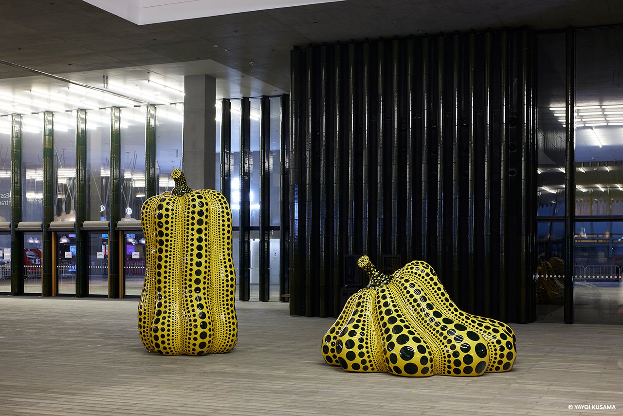 两个黄色南瓜雕塑并列摆放。右边的南瓜呈矮胖形，向左稍微下垂，左边的南瓜则呈细长形。两个南瓜的表面都满布黑色斑点，凸纹上的黑点比较大，凹槽内的黑点比较小。